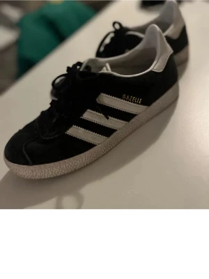 Svarta Adidas Gazelle sneakers - Säljer ett par klassiska Adidas Gazelle sneakers i svart mocka med vita detaljer och de ikoniska tre ränderna på sidan. Skorna har snörning, vit sula och Adidas-logga på hälen. Perfekta för dig som gillar retrostil och streetwear.
