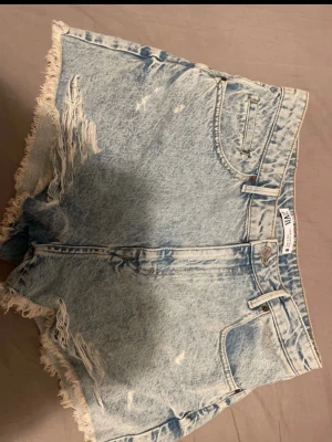 Ljusblå denimshorts från ZARA - Säljer ett par ljusblå denimshorts från ZARA med hög midja och råa, fransiga kanter. Shortsen har slitna detaljer framtill, klassiska fem fickor och knappgylf. Aldrig använt innan så de är även i perfekt skick. Perfekta för sommaren och ger en avslappnad vibe.🌸