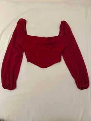 Röd korsettopp med puffärmar - Trendig röd korsettopp med långa puffärmar och fyrkantig urringning. Toppen har markerade sömmar framtill och dragkedja i ryggen för enkel påklädning. Snygg croppad passform som ger en edgy look.