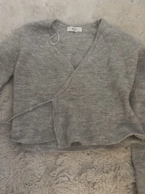 Grå omlottkofta från Gina Tricot - Mysig grå omlottkofta från Gina Tricot i ullblandning. Koftan har långa ärmar, knytband i sidan och en avslappnad passform. Perfekt för lager på lager under kyliga dagar.