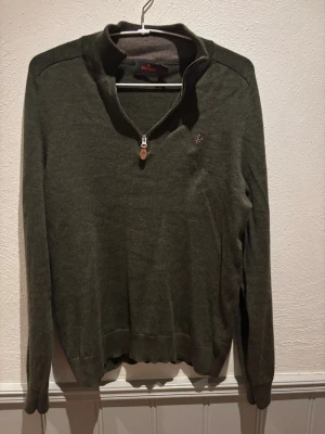 Olivgrön merinoullströja från Morris - Olivgrön långärmad tröja från Morris i 100% merinoull. Tröjan har en half zip-dragkedja vid halsen och en broderad logga på bröstet. Perfekt för lager-på-lagerr och snygg till jeans eller chinos. Storlek Xs. Har en liten defekt på insidan (se bild) men inget som stör eller gör varan något negativt. Nypris 1799.