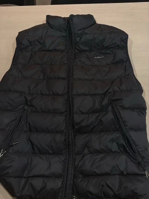 Svart dunväst från Gant XL - Säljer en svart dunväst från Gant i storlek XL. Västen har quiltad design, hög krage och dragkedja framtill samt två fickor med dragkedja. Perfekt att slänga över en hoodie eller tröja när det är kyligt ute.