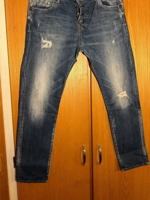  Replay jeans med slitningarblå denim - Säljer ett par blå jeans från Replay med snygga slitningar och tvättade detaljer. Klassisk femficksmodell med knäppning fram och kontrastsömmar. Jeansen har en rak passform och är tillverkade i tåligt denimtyg. Perfekta för dig som gillar en avslappnad och trendig look.