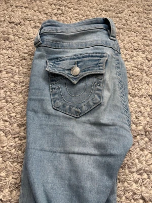 True Religion ljusblå bootcut jeans - Ljusblå bootcut jeans från True Religion i modellen Becca Mid Rise. Klassisk femficksdesign med ikonisk ficka bak och snygga kontrastsömmar. Jeansen är i mjukt denimtyg. Jeansen är slitna längst ner 