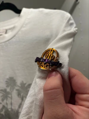 Vit Moncler x Palm Angels t-shirt - Säljer en clean vit t-shirt från Moncler i samarbete med Palm Angels. Framsidan har tryck med palmer och texten 'Snow Angels' i rosa. Klassisk rund hals och korta ärmar. På ärmen sitter en exklusiv guldfärgad Palm Angels x Moncler pin. Perfekt till streetwear-stilen. Liten fläck fram.