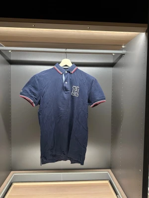 Marinblå piké från Tommy Hilfiger - Säljer en marinblå pikétröja från Tommy Hilfiger med vit och röd rand på krage och ärmslut. Pikén har en stor broderad H på bröstet och klassisk krage med knappar. Tillverkad i mjuk bomull, perfekt för en clean och sportig look.