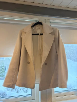 Beige kappa från H&M - Stilren beige kappa från H&M med klassisk krage och två knappar framtill. Jackan har raka ärmar, diskreta fickor och en mjuk, slät yta. Perfekt för dig som gillar en enkel men elegant look under höstiga/våriga dagar. Köpt för ca 500kr