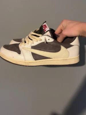 Nike Air Jordan 1 Low Travis Scott - Säljer ett par Nike Air Jordan 1 Low Travis Scott med ikonisk omvänd swoosh. Skorna har en kombination av brunt och vitt i mocka och skinn, med beige snörning och röda detaljer på hälen. Kommer med originalbox och specialdesignat rosa papper.