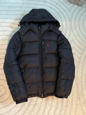 Svart pufferjacka från Polo Ralph Lauren - Svart pufferjacka från Polo Ralph Lauren med huva och röd logga. Jätte bra skick ser helt orörd ut vilket den nästan är! Skriv för fler frågor.