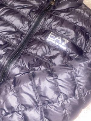 Svart pufferjacka från Emporio Armani - Säljer en svart pufferjacka från Emporio Armani EA7 med quiltad design och hög krage. Jackan har dragkedja framtill och logga på bröstet. Perfekt för dig som vill ha en stilren och varm jacka med snygg passform.