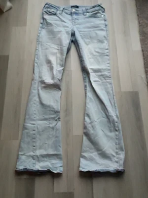 True Religion ljusblå bootcut jeans - Säljer ett par ljusblå jeans från True Religion med klassiska broderade fickor bak och bootcut-ben. Jeansen har låg midja och är tillverkade i mjukt bomullsmaterial. Snygga detaljer med knappar på fickorna och ikonisk True Religion-lapp bak.
