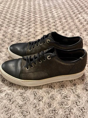 Lanvin DBB1 - Säljer ett par svarta sneakers från Lanvin med coolt strukturmönster på sidorna och tå i slätt skinn. Skorna har vita sulor, svarta skosnören och metallfärgade öljetter. Perfekta för dig som gillar stilrena men ändå unika detaljer.