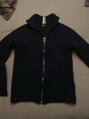 Mörkblå kofta från BOSS med dragkedja - Säljer en mörkblå kofta från BOSS i storlek xs Koftan har hel dragkedja framtill, ribbad krage med vita och beige detaljer samt diskret struktur i tyget. Perfekt för dig som gillar stilrena och klassiska plagg.