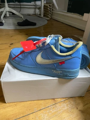 Nike Air Force 1 x Off-White blå - Säljer ett par Nike Air Force 1 x Off-White i blått skinn med silverfärgad swoosh och vit text på sidan. Skorna har vit snörning, synlig skumkant i gult och Off-White tag i rött. Klassisk perforerad tå och platt sula. Riktigt ikoniska sneakers för dig som vill sticka ut. Kommer också med extra laces. 