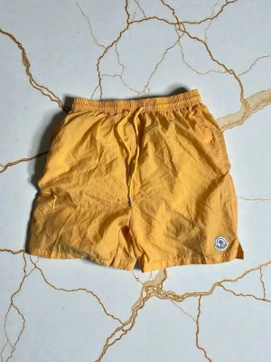 Gul Moncler badshorts med dragsko - Snygga gula badshorts från Moncler med elastisk midja och justerbar dragsko. Shortsen har sidofickor och en bakficka med knapp, samt Moncler-logga på vänster ben. Perfekta för strand eller poolhäng.