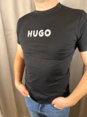 Svart HUGO BOSS t-shirt med logga - Snygg svart t-shirt från HUGO BOSS med stor HUGO-logga i vitt på bröstet. Klassisk rund halsringning och korta ärmar. Tillverkad i mjuk bomull som känns skön mot huden. Perfekt för dig som gillar stilrena och moderna plagg.