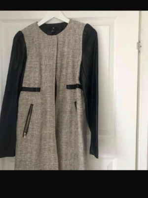 Beige kappa med svarta skinnärmar - Snygg beige kappa från H&M med svarta långa skinnärmar och svarta detaljer vid fickorna. Rak passform och dragkedjor på fickorna ger en modern look. Perfekt för dig som vill ha en stilren och trendig jacka.