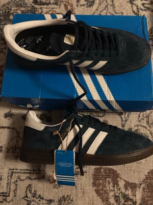 Adidas Handball Spezial eu44 - Adidas Handball Spezial sneakers i mörkblå mocka med vita detaljer och klassiska tre ränder på sidan. Skorna har brun gummisula, snörning och guldfärgad Spezial-logga. Insidan är svart med Adidas-tryck och hälen är vit. Perfekt för dig som gillar retrostil. Storlek 42 men sitter som 43/44!
