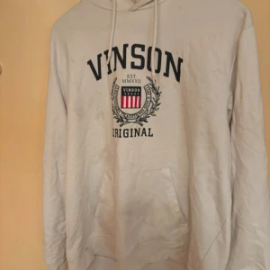 Vit hoodie från Vinson - Säljer en vit hoodie från Vinson med tryck framtill. Den har en klassisk design med dragsko i huvan och en stor ficka på magen. Perfekt för en avslappnad stil.