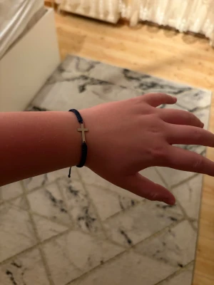 Blått armband med silverfärgat kors - Snyggt armband med mörkblå flätad tråd och ett stilrent silverfärgat kors i mitten. Justerbar knytning baktill gör att det passar de flesta handleder. Perfekt accessoar för dig som gillar enkel men betydelsefull design.