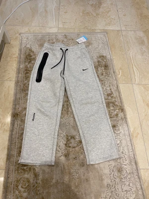 Nike NOCTA grå mjukisbyxor M - Snygga grå mjukisbyxor från Nike NOCTA med svart snörning i midjan och cool svart zip-detalj på ena benet. Byxorna har tryckta loggor och raka ben för en chill vibe. Perfekt för dig som gillar streetwear och vill ha något bekvämt men ändå stilrent.