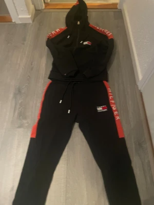 Svarta tracksuit Tommy Hilfiger - Svarta tracksuit från Tommy Hilfiger med röda sidopaneler och vit logotyptryck på benet. Byxorna har snörning i midjan och en slim passform. Perfekt för chill eller sportiga dagar, med mjukt material och snygg design.