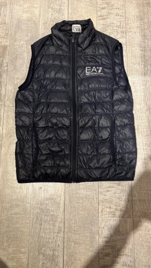Mörkblå dunväst från EA7 Emporio Armani - Snygg mörkblå dunväst från EA7 Emporio Armani med quiltad design och dragkedja framtill. Västen har hög krage och logga på bröstet. Perfekt för lager-på-lager och streetstyle. Materialet är glansigt och lätt, vilket ger en modern look.