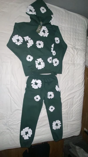 Grön hoodie med vita blommor - Mörkgrön hoodie med huva och stora vita blommiga tryck över hela tröjan. Hoodien har en avslappnad passform och är tillverkad i mjuk bomull, perfekt för chill dagar. Snyggt och unikt mönster som sticker ut. Matcha med de medföljande byxorna för en komplett look. 10/10 i skick och den är oanvänd, finns att köpas separat men billigare att köpa hela setet
