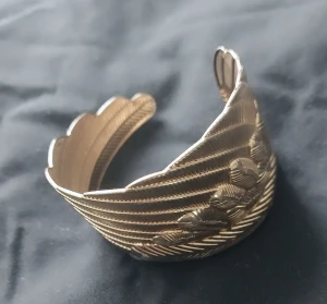 Guldfärgat armband i fjäderdesign - Unikt guldfärgat armband med snygg fjäderform och detaljerade linjer som ger en cool struktur. Armbandet är öppet och har en bred, markerad design som verkligen sticker ut. Perfekt accessoar för dig som vill ha något extra till din stil.