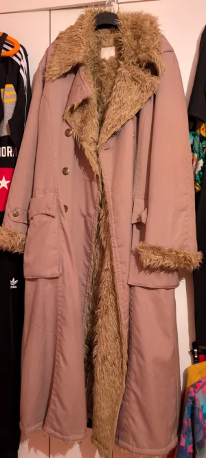 Beige pälsjacka med stora fickor - Säljer en lång beige pälsjacka med fluffig fuskpäls på krage, insida och ärmslut. Jackan har stora fickor med lock, snygga knappar och en loose passform. Perfekt för dig som vill ha en unik och varm look i vinter. Passar både Medium och Large.Det är nog en herrrock från början lik en sk "lottarock" .Bra skick behöver dock vädras ordentligt. 