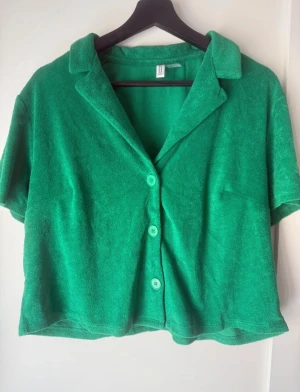 Grön frottéskjorta med kort ärm - Snygg grön skjorta från Monki i mjuk frotté med korta ärmar och klassisk krage. Skjortan har tre gröna knappar framtill och en avslappnad, loose passform. Perfekt för dig som gillar färg och vill ha något bekvämt och chill.