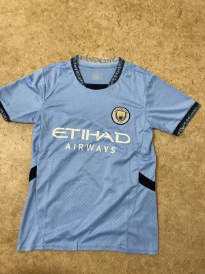 Manchester City ljusblå fotbollströja - Säljer en officiell Manchester City fotbollströja i ljusblått med subtilt geometriskt mönster. Tröjan har korta ärmar, svart detalj vid ärmslut och krage, samt klubbmärke och Etihad Airways-tryck på bröstet. Tillverkad i lätt och ventilerande polyester.