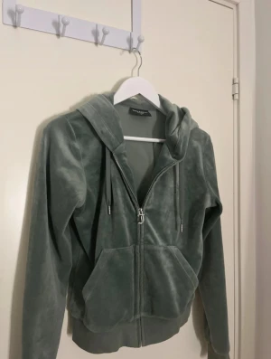 Grön velour hoodie Juicy Couture M - Mjuk grön hoodie från Juicy Couture i velour med dragkedja och klassisk Juicy J-detalj på zippen. Tröjan har huva, kängurufickor och ribbade muddar. Perfekt för en chill och trendig look. köpt för 1200 men andvänds inte mer och därför säljs den.🩷