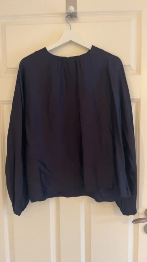 Nu mörkblå blus i satin  - Säljer en helt ny mörkblå blus i satin med rund halsringning. Blusen har en liten öppning med knapp i nacken och en lätt glansig finish som ger en lyxig känsla. Perfekt för dig som gillar stilrena och enkla plagg. Blusen är från WERA och säljs inte längre 