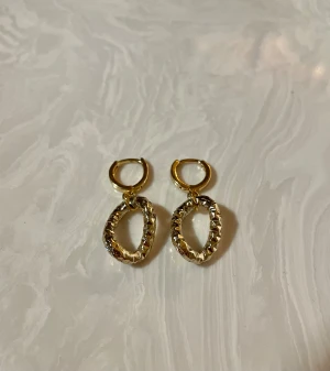 Guldiga ovala örhängen med struktur - Säljer ett par snygga örhängen i guld med oval form och coolt strukturerad yta. De har små ringar upptill och hängande ovala delar som ger en trendig vibe. Perfekta för att lyfta vilken outfit som helst.