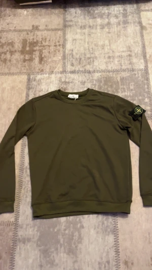 Olivgrön Stone Island sweatshirt L - Snygg olivgrön sweatshirt från Stone Island i storlek L. Tröjan har klassisk rund hals och lång ärm, med den ikoniska Stone Island patchen på vänster ärm. Tillverkad i mjuk bomull som är skön mot huden och passar perfekt till streetwear-stilen.