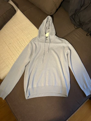 Ljusblå stickad hoodie från Bvaldi - Snygg ljusblå hoodie från Bvaldi med huva och snörning. Tröjan är stickad i mjukt material och har ribbade muddar vid ärmslut och nederkant. Liten broderad logga på bröstet där det står bvaldi. Inte använd en ändå gång, var lite för liten, modellen är 182 cm. 