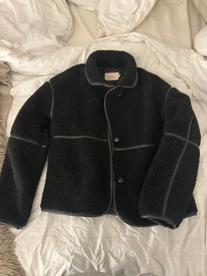 Svart teddyjacka från Nelly - Säljer en svart teddyjacka från Nelly med snygga detaljer i skinnimitation längs sömmarna. knappar framtill, fluffigt material som håller dig varm. Perfekt för dig som gillar stilrena och mysiga plagg. Har aldrig använts 