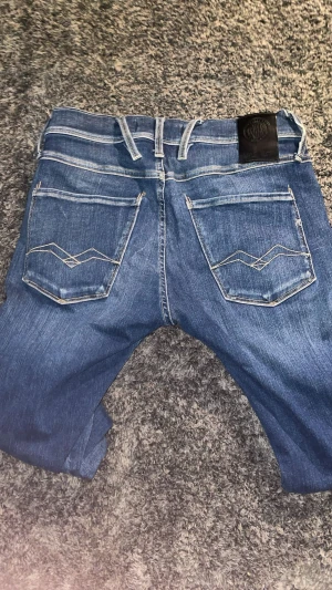 Replay Anbass blå jeans, storlek 28 - Snygga Replay Anbass jeans i klassisk blå tvätt med slitningar och kontrastsömmar. Modellen har fem fickor, smal passform och tydliga detaljer på bakfickorna. Perfekta för dig som gillar en modern och stilren look. Pris kan diskuteras och storleken är S