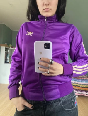 Lila Adidas vindjacka  - Säljer en lila Adidas vindjacka i storlek S med klassiska vita och gula stripes längs ärmarna. Jackan har dragkedja framtill, ribbade muddar och ståkrage. Adidas-logga på bröstet och backsidan, och fickor med dragkedja. Perfekt för en sportig och chill look.