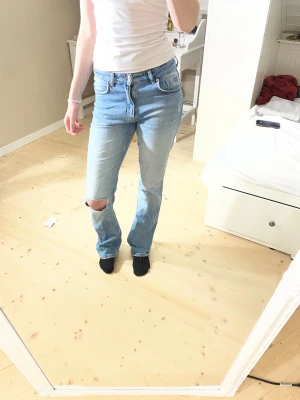 Ljusblå jeans från Gina Tricot - Säljer ett par ljusblå jeans från Gina Tricot i storlek 38. Modellen har utsvängda ben, klassisk femficksdesign och en snygg slitning på ena knät. Jeansen har hög midja och är tillverkade i bomull med gul kontrastsöm. Liten defekt på höger bakficka. Modellen är 172cm lång.