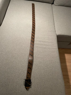 Fendi bälte i skinn med FF-logga - Säljer ett snyggt Fendi-bälte i svart och brunt skinn med den klassiska FF-loggan i hela längden. Bältet har en svart metallspänne formad som Fendi-loggan och är vändbart med en helsvart sida. Perfekt accessoar för att lyfta din outfit.