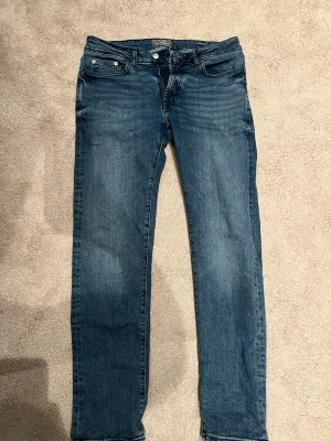 Blå jeans från Dressmann, storlek 30/30 - Snygga blå jeans från Dressmann i klassisk slim fit. Jeansen har fem fickor, knapp och dragkedja framtill samt diskret tvättad look. Perfekt passform för dig som gillar en normal siluett och tidlös stil.