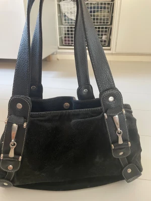 Svart axelväska i mocka och skinn - Snygg svart axelväska med breda handtag i skinn och kropp i mjuk mocka. Väskan har silverfärgade metalldetaljer och flera fack med dragkedja inuti. Köpt second hand och använt många gånger ☺️ 