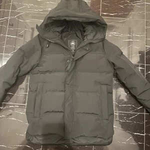 Svart canada goose black label jacka  - Svart pufferjacka från Canada Goose med huva och broderad logotyp på ärmen. Jackan har dragkedja och tryckknappar framtill, två sidofickor och quiltad design. Perfekt för kalla dagar med sin tjocka och isolerande fyllning.