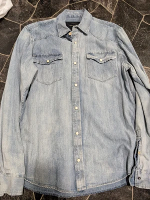 Ljusblå jeansskjorta Skill Casual Wear stl M - Snygg ljusblå jeansskjorta från Skill Casual Wear med klassisk western-look. Skjortan har två bröstfickor med lock och tryckknappar framtill. Tillverkad i bomull och har långa ärmar. Perfekt för en avslappnad och cool stil.