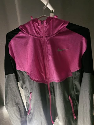 Nike vindjacka ( rosa & grå )  - Nike vindjacka i snygg rosa och grå färg med svarta detaljer. Jackan har huva, hel dragkedja och samt två fickor framtill. Lätt och tunn i polyester, perfekt för löpning eller träning. Reflekterande Nike-logga på bröstet och har en väldigt sportig passform.                                       OBS! Stor i storlek, passar även storlek M. 👍