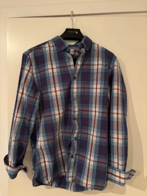 Blårutig skjorta från Dobber  - Snygg blårutig skjorta från Dobber i storlek medium. Skjortan har klassisk krage, långa ärmar och knappar framtill. Färgerna är blå, vit och röd i ett rutigt mönster