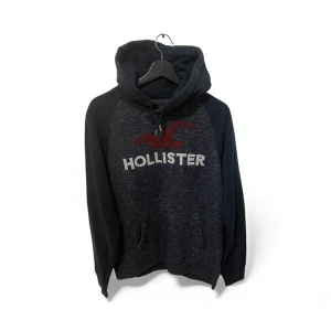 Mörkblå hoodie från Hollister - Snygg mörkblå hoodie från Hollister med stor logga och röd fågel på bröstet. Tröjan har huva med snörning, känguruficka och långa ärmar. Perfekt för chill dagar och har en skön, mjuk känsla tack vare bomullsmaterialet.