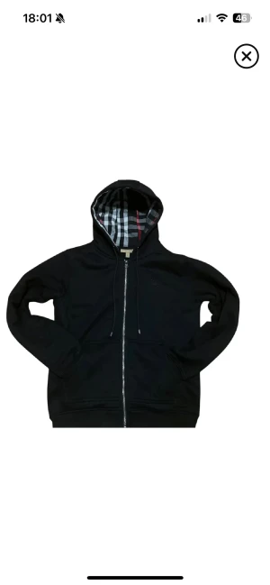 Svart Burberry hoodie med dragkedja - Svart hoodie från Burberry med klassiskt rutigt foder i huvan och längs insidan. Tröjan har hel dragkedja, snörning i huvan och två fickor framtill. Perfekt för dig som gillar stilrena och exklusiva streetwear-plagg.